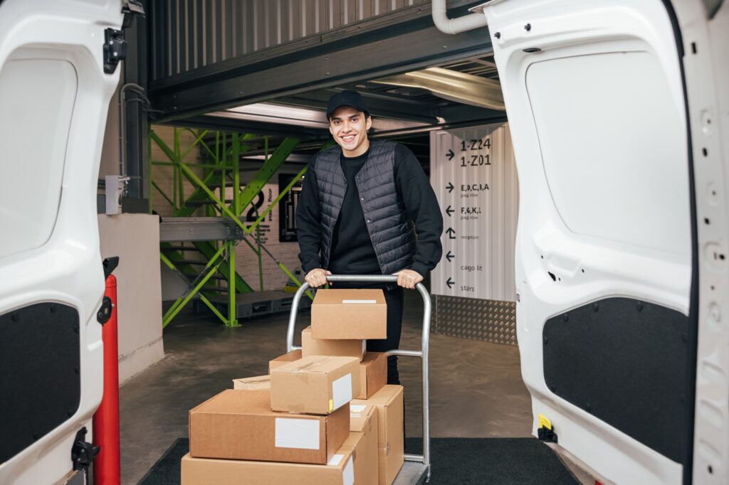 Smiling courier in uniform standing in warehouse with cart preparing for delivery